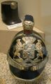 Helm van de Marechaussee in gebruik van 1844 tot 1869. Op de voorkant het koninklijk wapen van 1815/’16. Links boven de officierssjako model 1860. (Coll. Mus. Kon. Marechaussee, Buren)